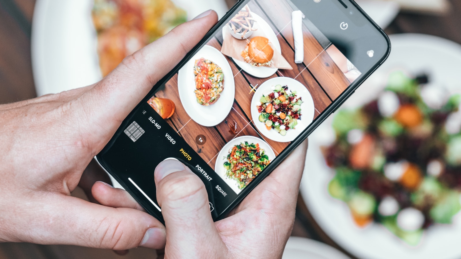 Hände fotografieren mit einem Smartphone angerichtete Speisen auf einem Restauranttisch.